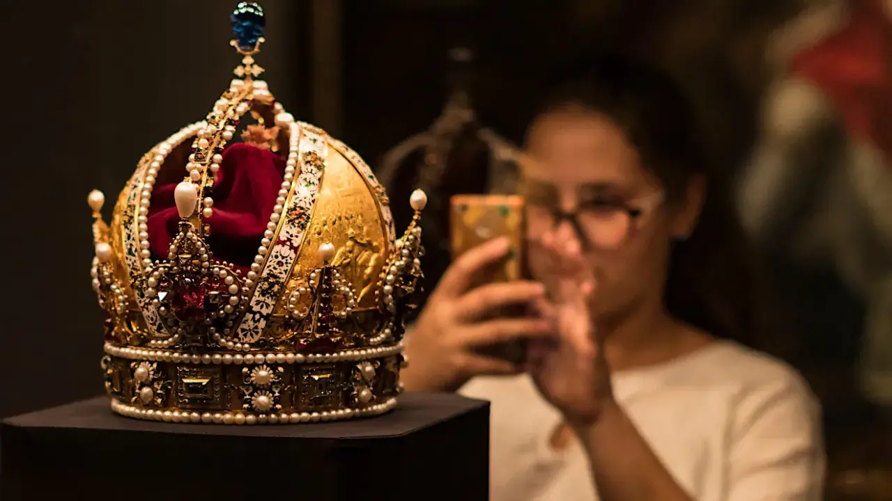 Złota korona, ozdobiona perłami i rubinami, przypomina wielki mogoł diament. Kobieta w tle robi jej zdjęcie telefonem.