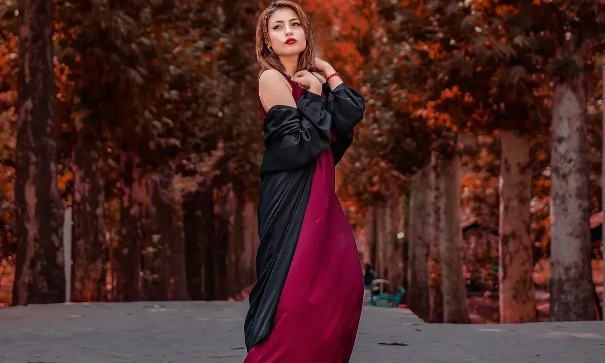 Elegancka kobieta w bordowej sukience i czarnej narzutce, z subtelną biżuterią, pozuje na tle jesiennej alei.