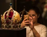 Złota korona, ozdobiona perłami i rubinami, przypomina wielki mogoł diament. Kobieta w tle robi jej zdjęcie telefonem.