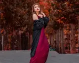 Elegancka kobieta w bordowej sukience i czarnej narzutce, z subtelną biżuterią, pozuje na tle jesiennej alei.
