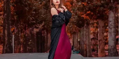 Elegancka kobieta w bordowej sukience i czarnej narzutce, z subtelną biżuterią, pozuje na tle jesiennej alei.