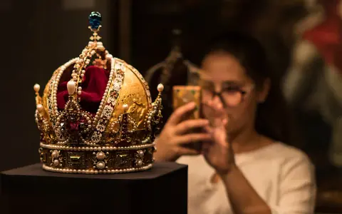 Złota korona, ozdobiona perłami i rubinami, przypomina wielki mogoł diament. Kobieta w tle robi jej zdjęcie telefonem.