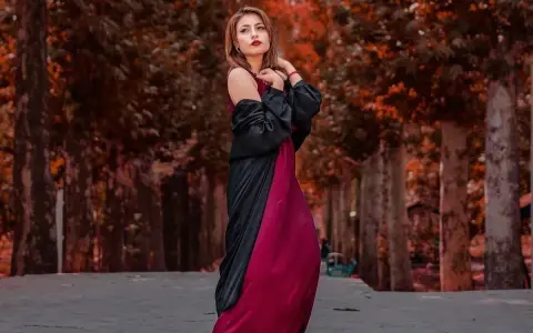 Elegancka kobieta w bordowej sukience i czarnej narzutce, z subtelną biżuterią, pozuje na tle jesiennej alei.