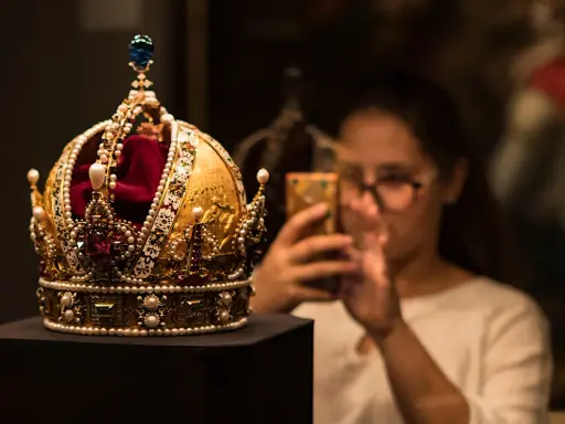Złota korona, ozdobiona perłami i rubinami, przypomina wielki mogoł diament. Kobieta w tle robi jej zdjęcie telefonem.