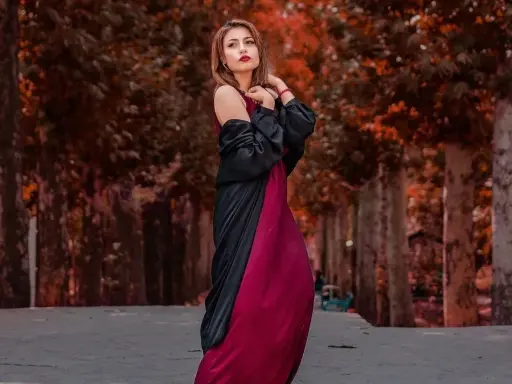 Elegancka kobieta w bordowej sukience i czarnej narzutce, z subtelną biżuterią, pozuje na tle jesiennej alei.