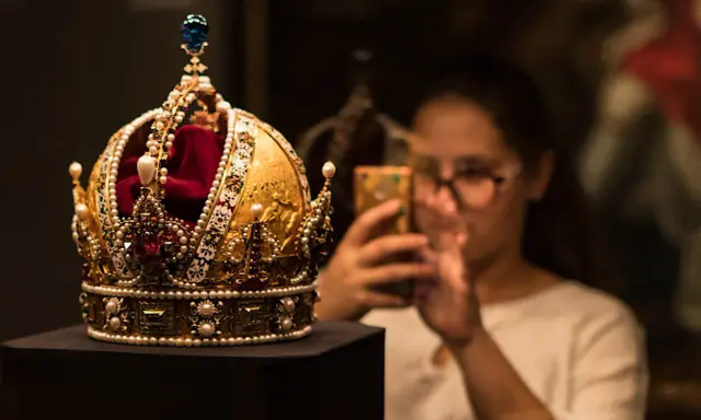 Złota korona, ozdobiona perłami i rubinami, przypomina wielki mogoł diament. Kobieta w tle robi jej zdjęcie telefonem.
