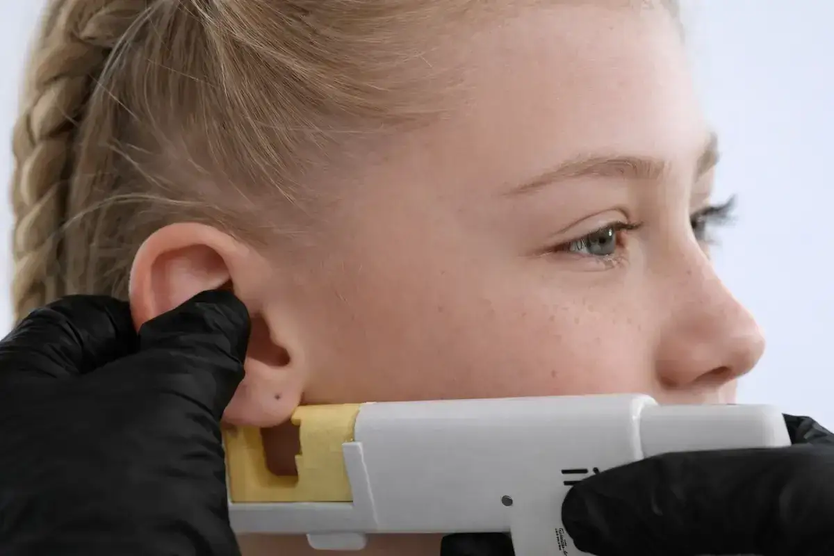 Ucho dziewczynki z kolczykiem, gdzie najbardziej boli kolczyk w uchu? Pistolet do przekłuwania uszu w rękach osoby w czarnej rękawiczce.