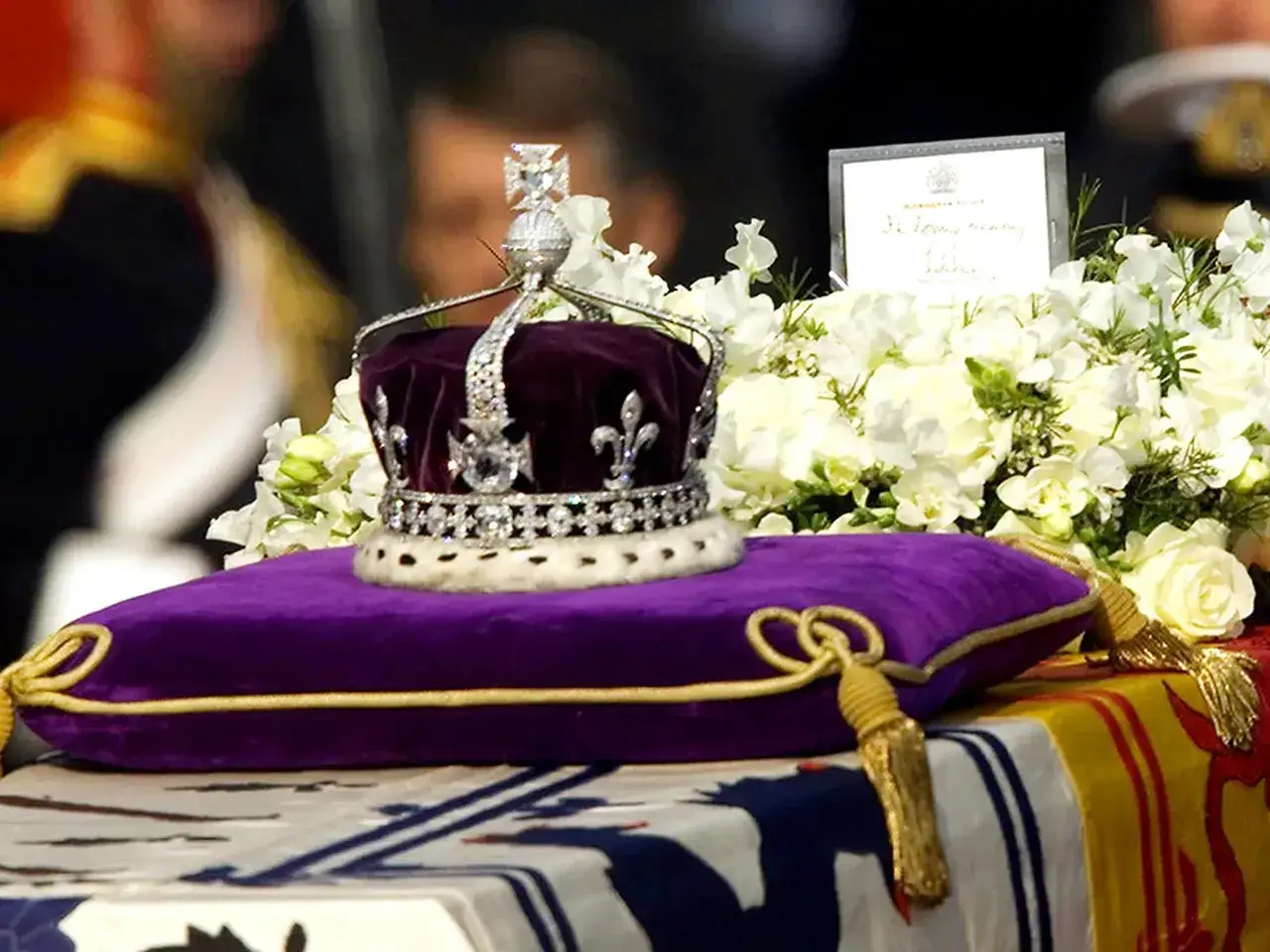 Korona z diamentem Kohinoor na aksamitnej poduszce, otoczona białymi kwiatami.